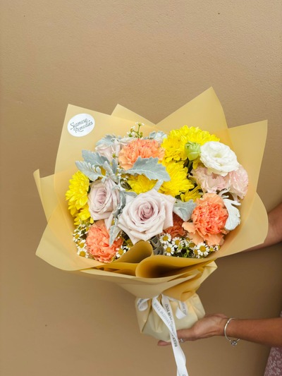 A cheerful bouquet with yellow chrysanthemums, blush roses, peach carnations, and white lisianthuses in golden wrapping.