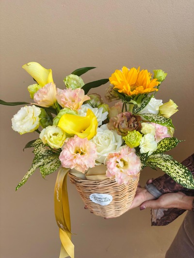 A woven basket filled with a sunflower, yellow calla lilies, lisianthus, roses, and greenery, finished with a golden ribbon.
