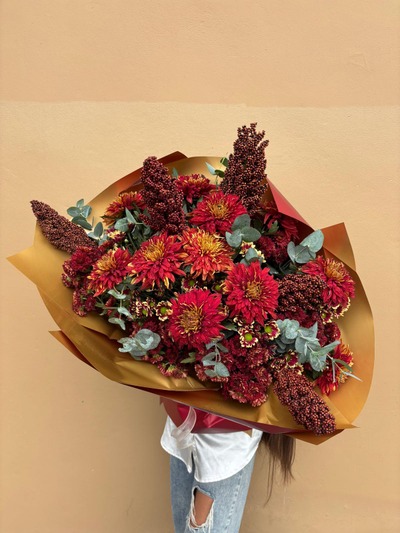 A bouquet of deep red chrysanthemums and textured blooms with eucalyptus, wrapped in golden paper.