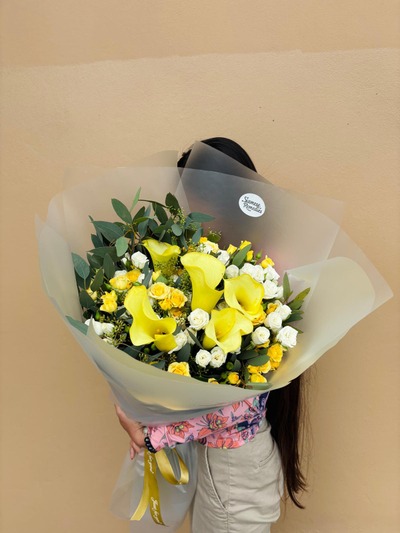 A radiant bouquet featuring yellow calla lilies, white roses, and yellow spray roses wrapped in soft paper.